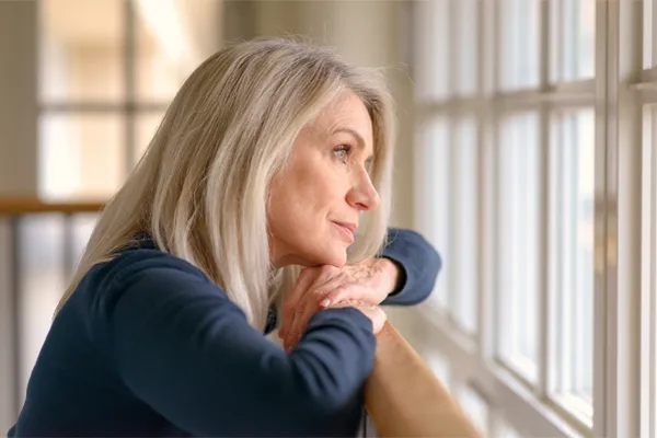 Adrenal-Hormone-Disorder-Clinic A mature woman in a blue shirt looks out the window with melancholy. Schedule treatment for adrenal hormone disorder from Morgan Burns, APRN in Central Florida.