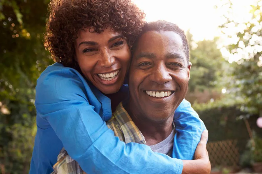 Autoimmune-Doctor A middle-aged African American couple outside, the woman embracing the man from behind pressing her cheek to his, both are smiling due to successful Autoimmune Disorder treatment from Morgan Burns, APRN in Central Florida.