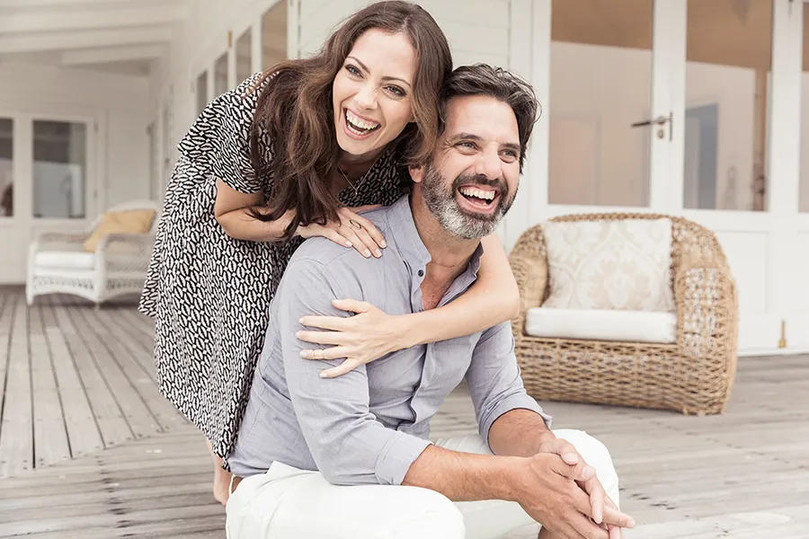 Concierge-Care A middle-aged couple on a wooden porch with wicker furniture; the woman is playfully hugging the man from behind. They are benefiting from the concierge care services from Morgan Burns, APRN in Central Florida.