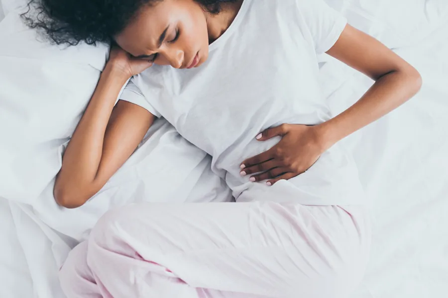 Crohns-Disease-Clinic A dark-skinned woman in white clothes lays on her bed clutching her middle before getting treatment for Crohn's Disease from Morgan Burns, APRN in Central Florida.