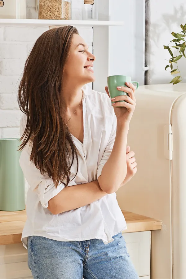 Crohns-Disease-Treatment A woman with a white blouse an jeans drinking tea in her kitchen, smiling after getting treatment for Crohn's Disease from Morgan Burns, APRN in Central Florida.