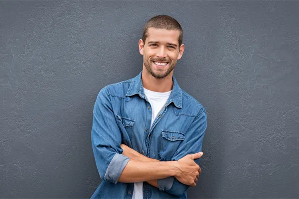Hormones-for-Men-Doctor A man in a denim shirt stands smiling against a gray-blue wall, pleased with his testosterone hormone therapy from Morgan Burns, APRN in Central Florida.