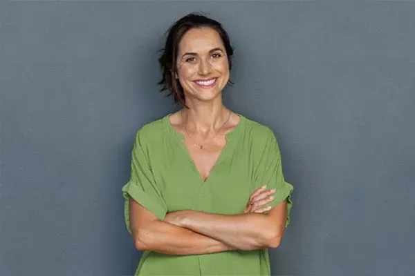Hormones-for-Women-Doctor A woman in a green shirt stands smiling against a gray-blue wall, pleased with her hormone therapy from Morgan Burns, APRN in Central Florida.
