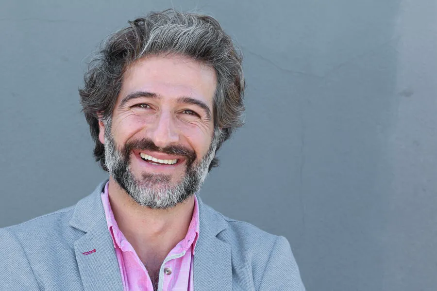 Hypogonadism-Clinic A smiling man with salt and pepper hair and beard wearing a pink shirt and gray blazer stands agains a gray wall. Get hypogonadism treatment from Morgan Burns, APRN in Central Florida.