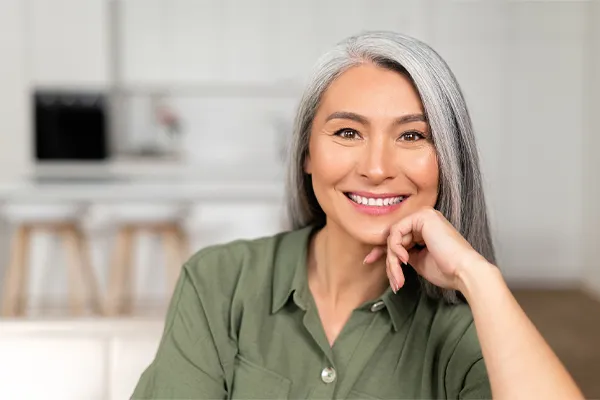 Menopause-Clinic An asian woman with long gray hair and a green shirt sitting and smiling on her couch, happy with her menopause care from [NAME} in Central Florida.