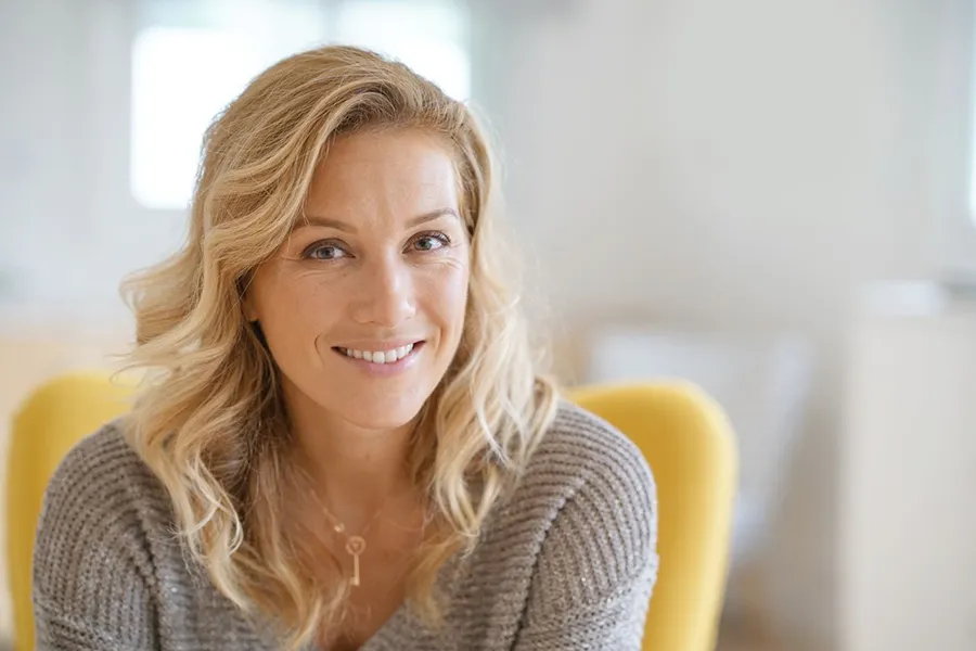 Perimenopause-Doctor A blonde woman wearing a a gray sweater sits inside in a yellow chair, smiling and happy with her perimenopause treatment from Morgan Burns, APRN in Central Florida.