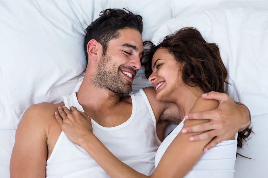 Sexual-Health-Peptide-Therapy-Doctor A laughing man and woman in undershirts lie in each other's arms on a white bed, representing sexual health peptide therapy by Morgan Burns, APRN in Central Florida.