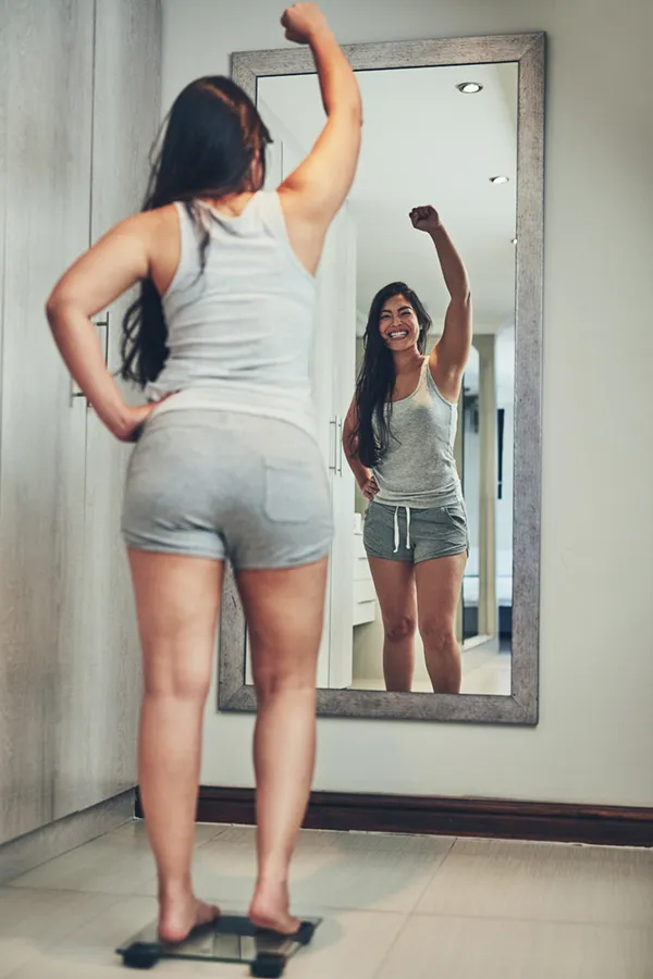 Tirzepatide-Treatment A woman in workout clothes stands on a scale looking in the mirror and pumping her hands in celebration of her successful Tirzepatide treatment from Morgan Burns, APRN in Central Florida.